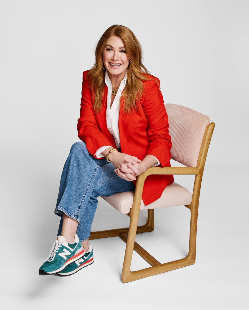 Dee, pH-D Founder, in a red blazer and blue jeans sitting on a chair against a white background