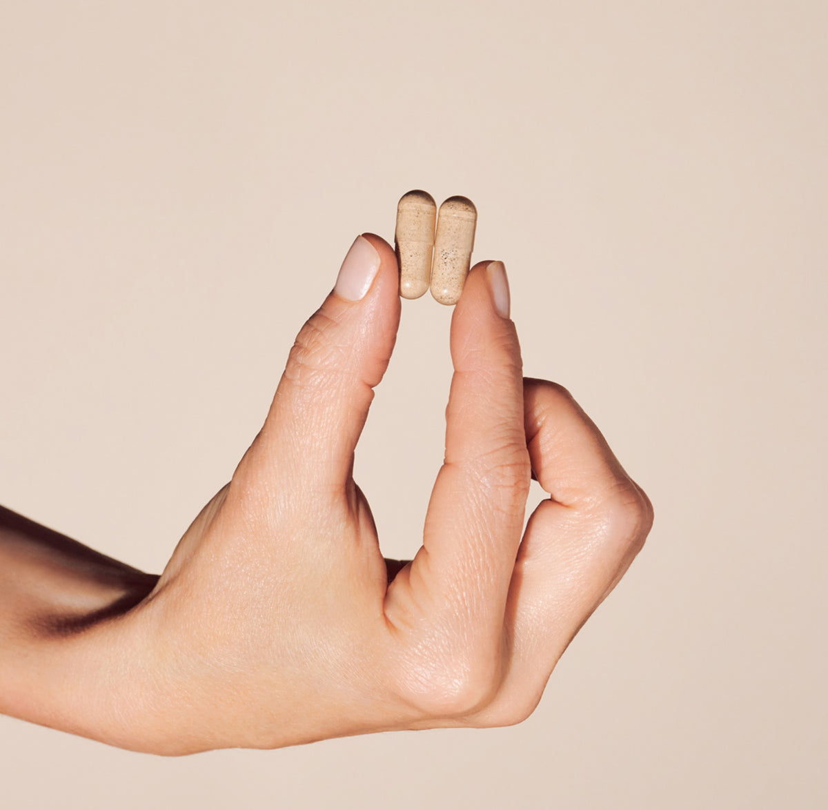 A close-up of a hand holding two small beige capsules between the fingers against a plain warm beige background.