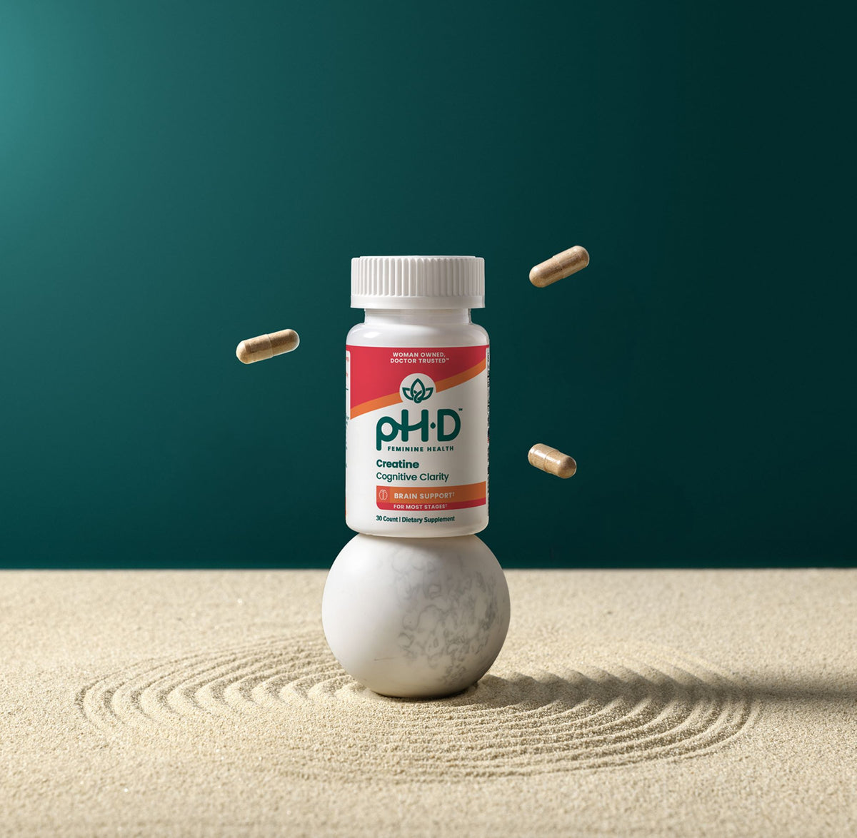 pH-D Creatine Cognitive Clarity stands on a white marble sphere in a styled studio setup. Several capsules float around the bottle against a dark teal background, with a light beige textured surface in the foreground showing concentric ripple lines.
