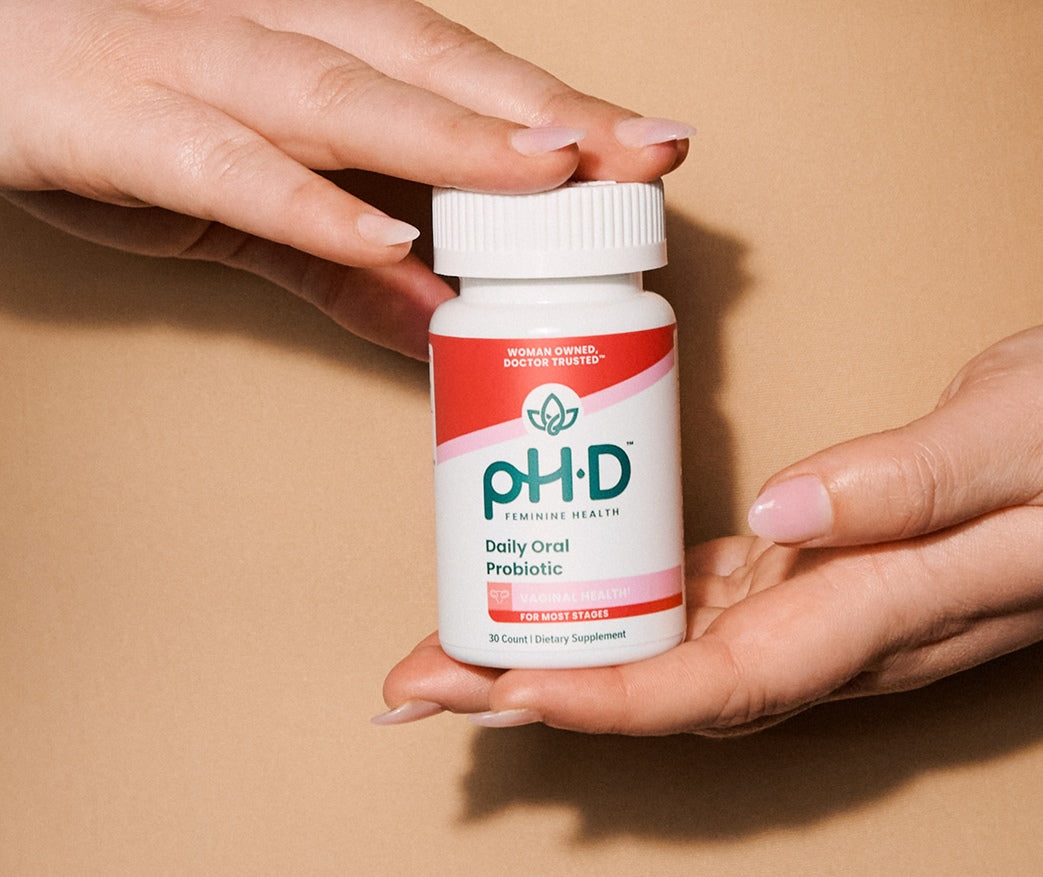 A white bottle of pH-D Feminine Health Probiotic held between two hands against a solid tan background.