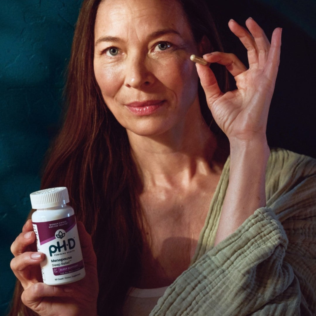 A woman holds a bottle of pH-D Menopause Sleep Relief in one hand and brings a single capsule to her eye with the other hand, set against a dark teal textured background.