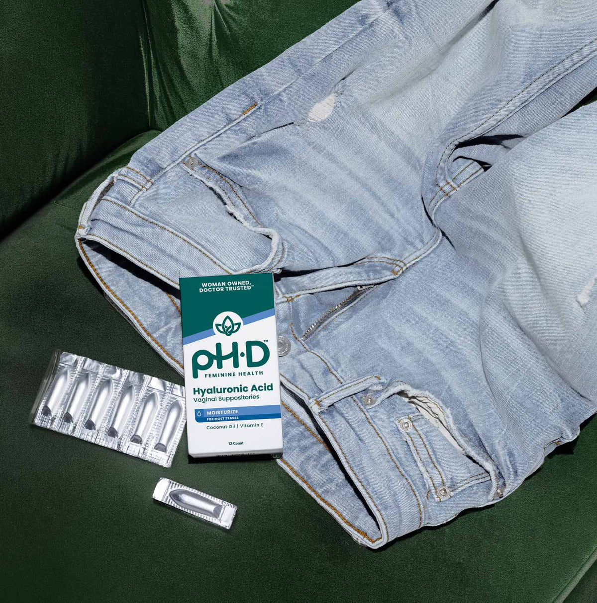 A pair of light blue distressed jeans laid on a dark green couch next to a box of pH-D Feminine Health Hyaluronic Acid vaginal suppositories and two silver blister packs