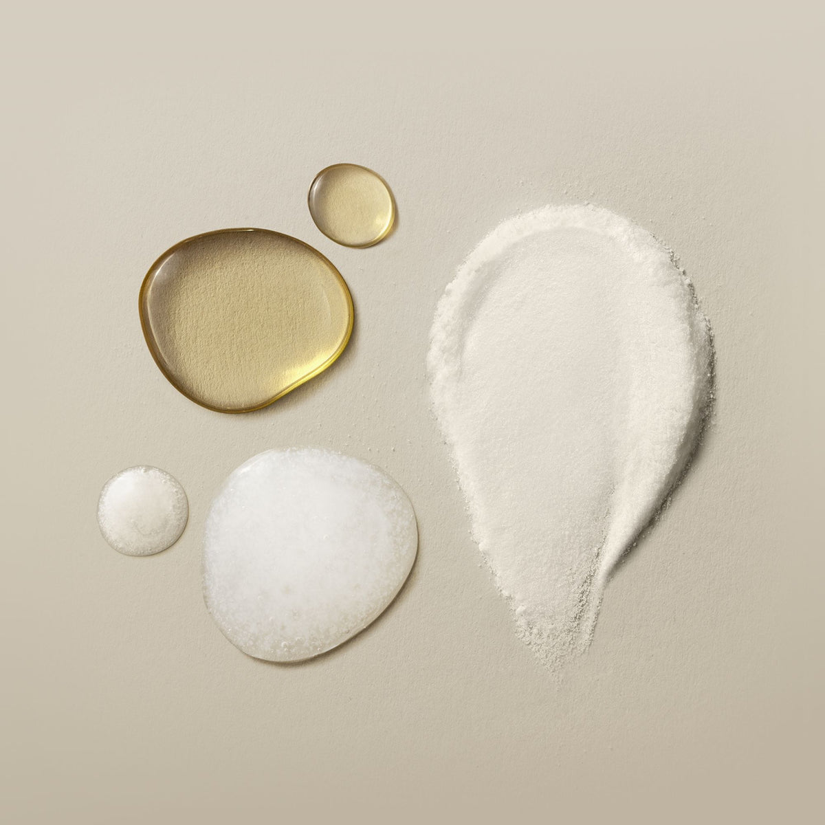 A cosmetic ingredient display on a warm beige background, featuring a large teardrop-shaped mound of white powder alongside two translucent golden liquid droplets and two small white gel or cream droplets.