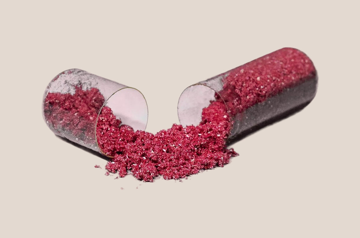 pH-D Feminine Health Probiotic capsule tipped over with sparkling dark pink powder spilled out onto a light beige background. 