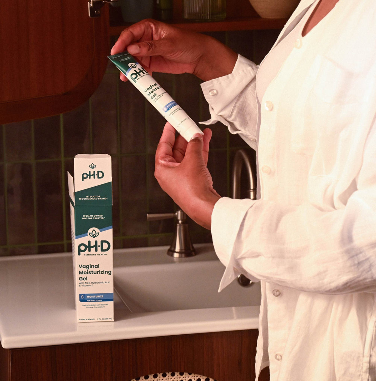 A person in a white shirt holds a tube of pH-D Feminine Health Vaginal Moisturizing next to its box on a bathroom sink counter.