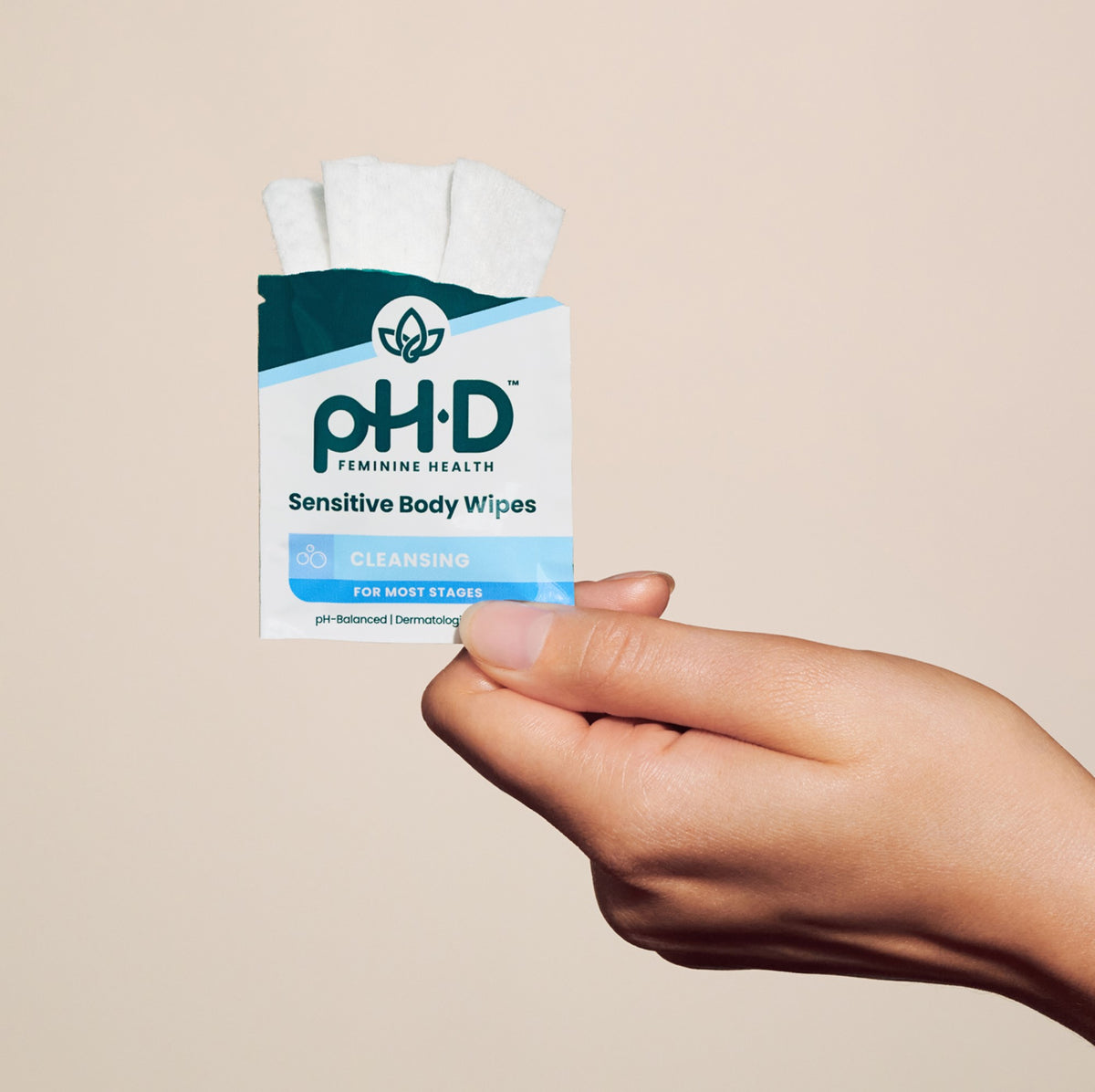 A hand holding a single packet of pH-D Feminine Health Sensitive Body Wipes against a solid beige background.