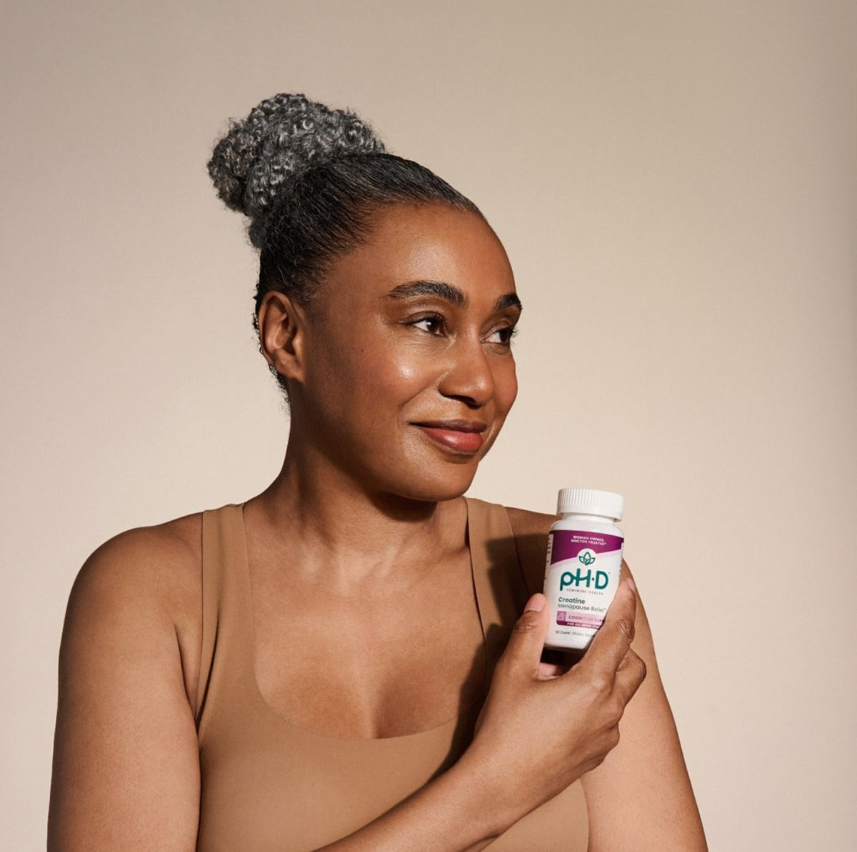 A woman with her hair in a high bun holds a bottle of pH-D Feminine Health Creatine Menopause Relief against a solid beige background.